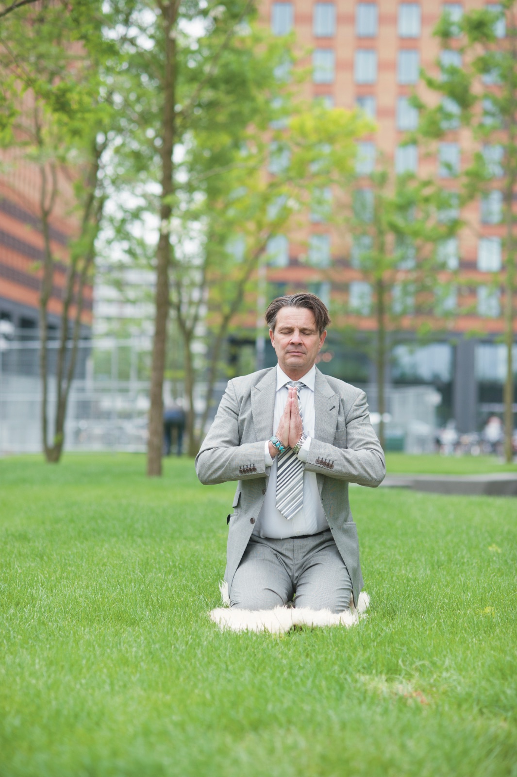 A man in a business suit meditating on a lawn between office buildings